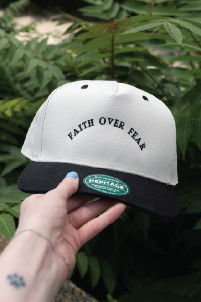 White and black snapback hat with the phrase ‘Faith Over Fear’ embroidered in black on the front, held up outdoors in front of green leafy plants.